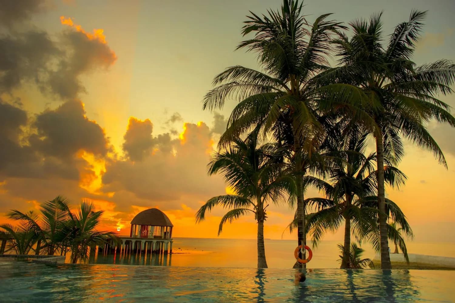 Malediven Sonnenuntergang mit Palmen, Überwasser-Pavillon und Infinity-Pool am Meer