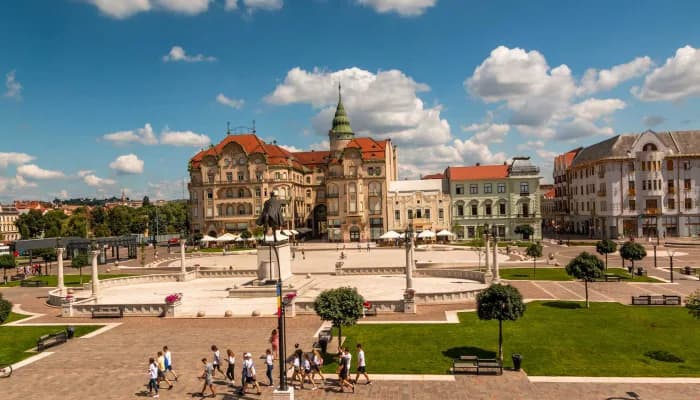 Großer Platz in Oradea mit Reiterstatue, Jugendstilgebäude, grünen Rasenflächen und Spaziergängern unter blauem Himmel