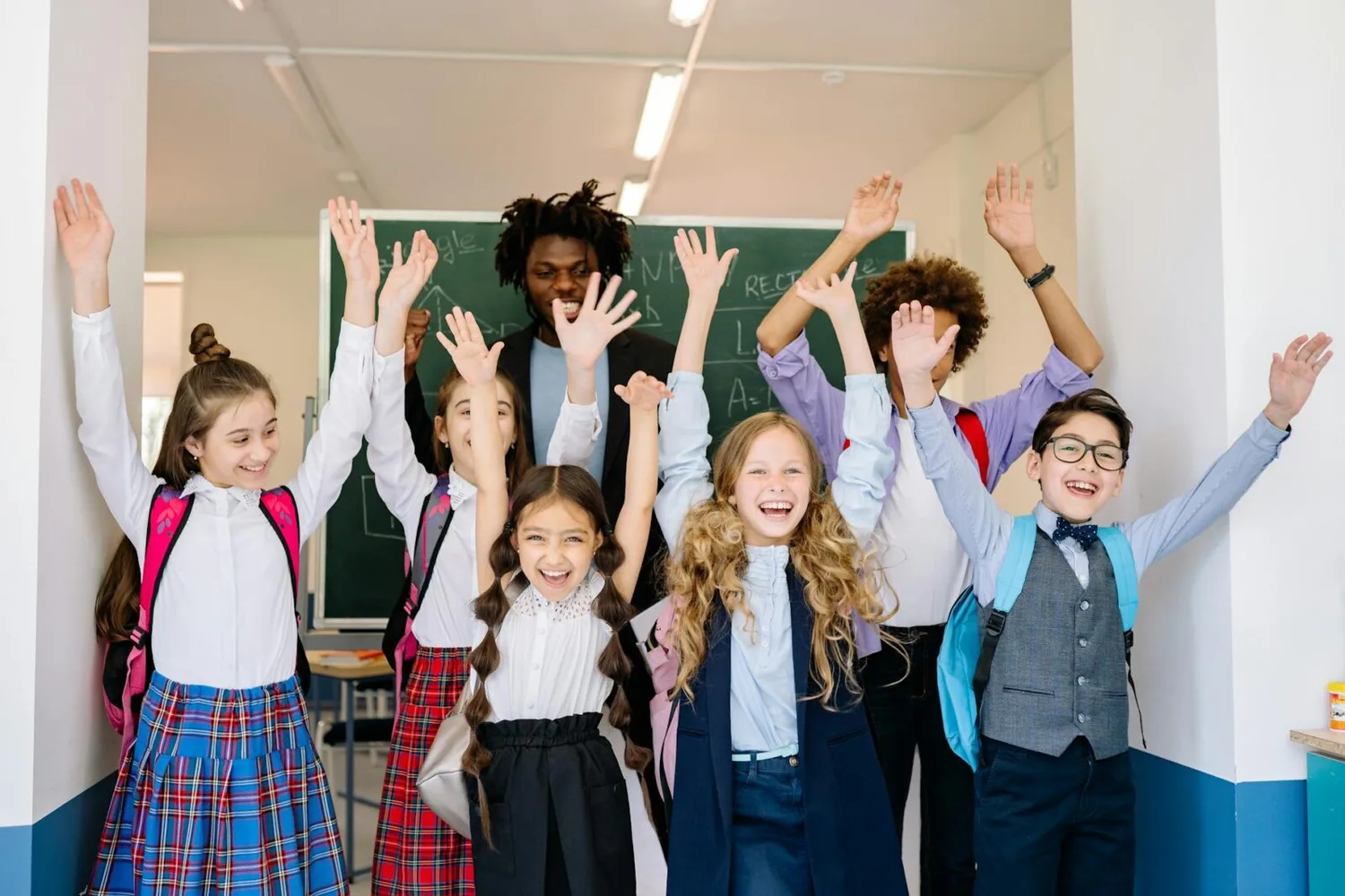 Schulklasse jubelt im Klassenzimmer mit Lehrkraft im Hintergrund, Kinder mit Rucksäcken feiern Teilnahme am Videowettbewerb Flughafen Hamburg