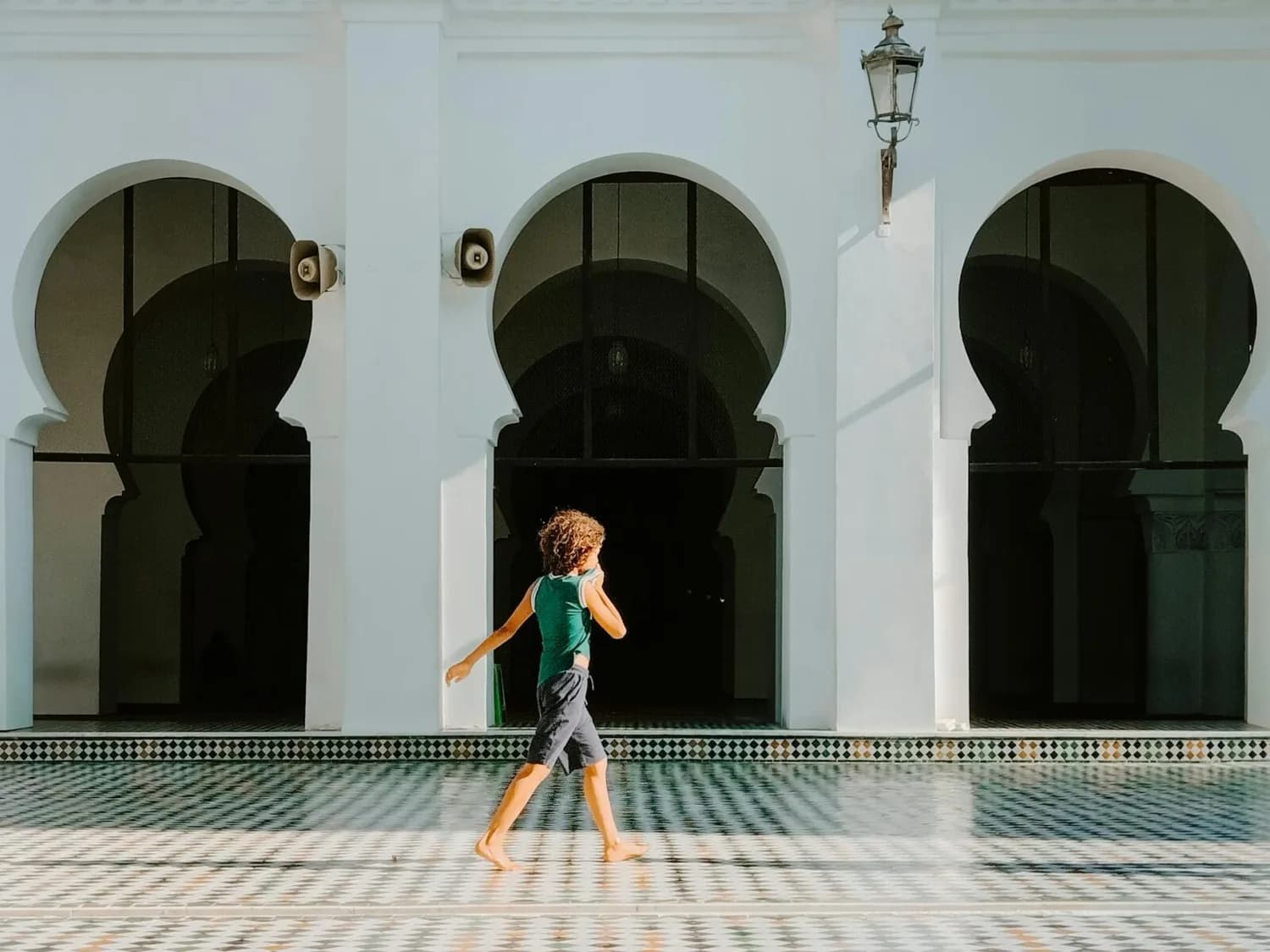 Person in leichter Sommerkleidung geht barfuß durch Innenhof mit gemustertem Fliesenboden vor weißen, maurisch inspirierten Arkaden in Fès, Marokko – günstiger Urlaubsszene.