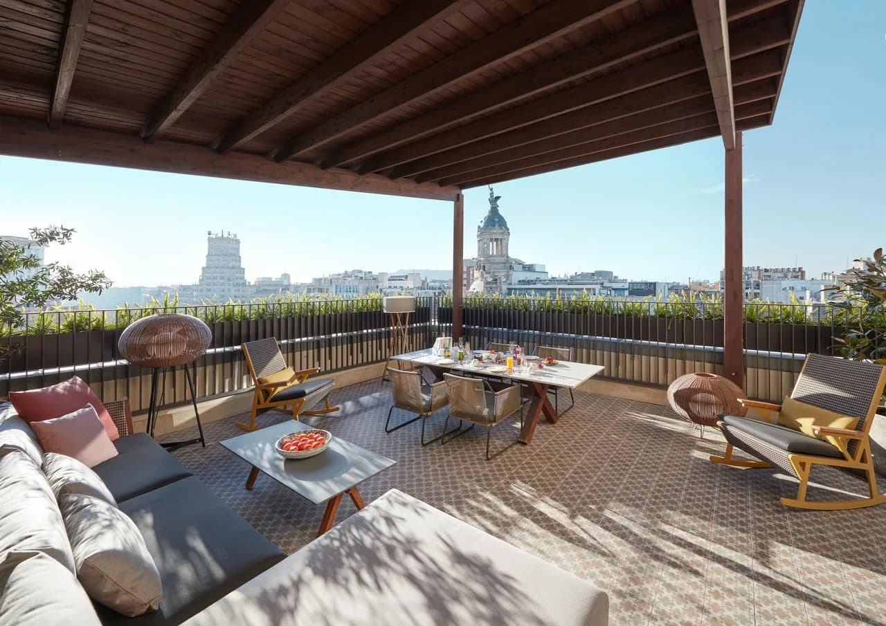 Penthouse-Terrasse der Mandarin Oriental Barcelona mit überdachtem Lounge- und Essbereich, Sonnenliegen, Tisch mit Frühstück und Blick auf die Stadt und historische Kuppel