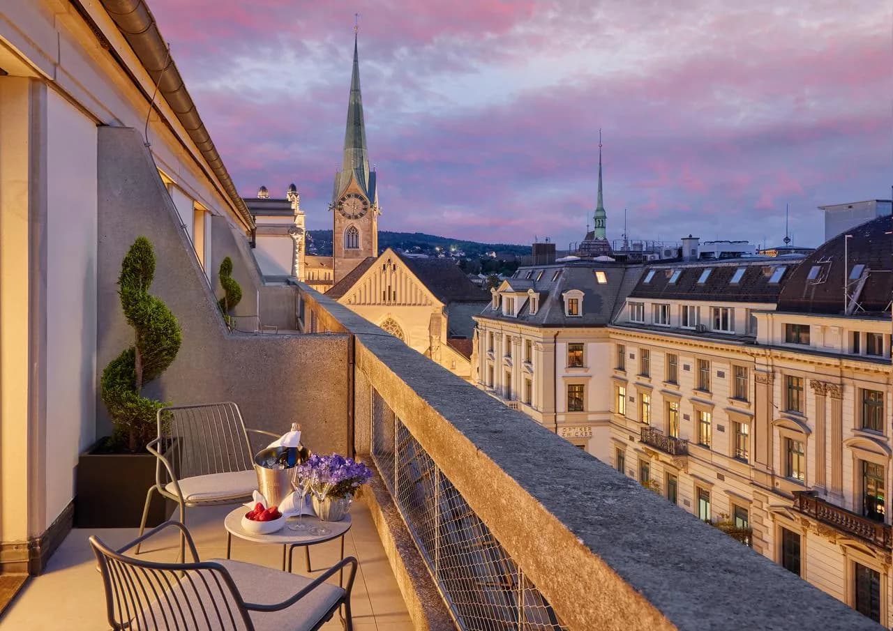 Terrassenblick vom Deluxe-Zimmer Mandarin Oriental Savoy Zürich mit Sitzbereich, Champagner auf Tisch und Blick auf historische Kirchen- und Stadthäuser bei Sonnenuntergang