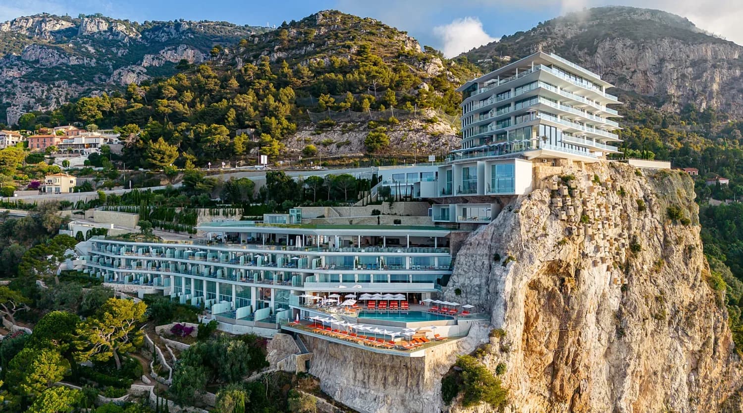 Luxuriöses Maybourne Riviera Hotel Cap Martin an steiler Felsküste mit Terrassenpools, Sonnenliegen und Panorama-Glasbalkonen vor mediterranen Bergen in Frankreich