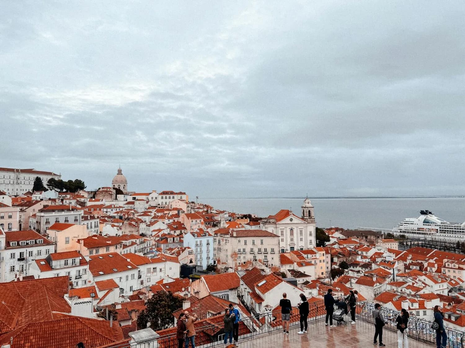 Panorama von Lissabon mit roten Dachhäusern, Alfama, Kirchenkuppel und Blick aufs Tejo-Flussufer, Besucher an Aussichtsterrasse