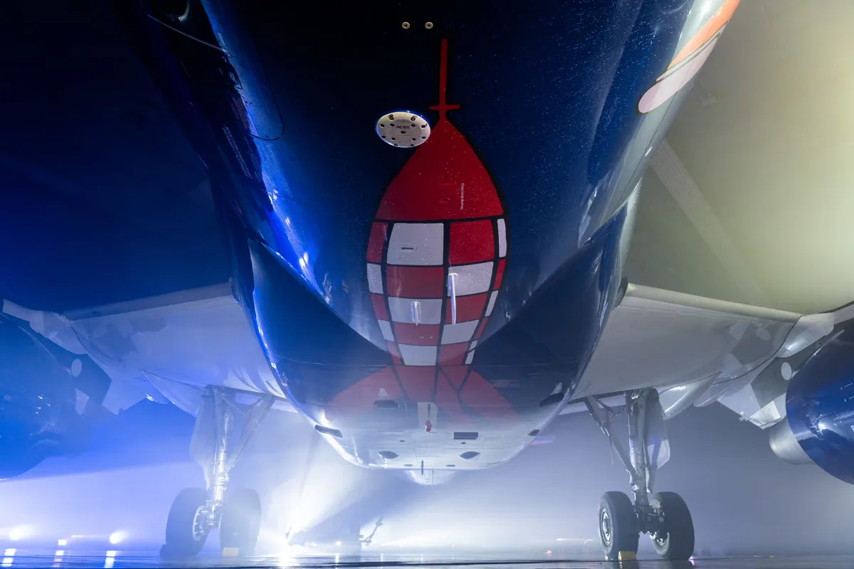 Unteransicht eines Brussels Airlines Passagierflugzeugs mit roter Raketen-Grafik am Rumpf, Fahrwerk ausgefahren und beleuchteter Hangar-Szenerie - © Brussels Airlines / Hergé / Tintinimaginatio 2026
