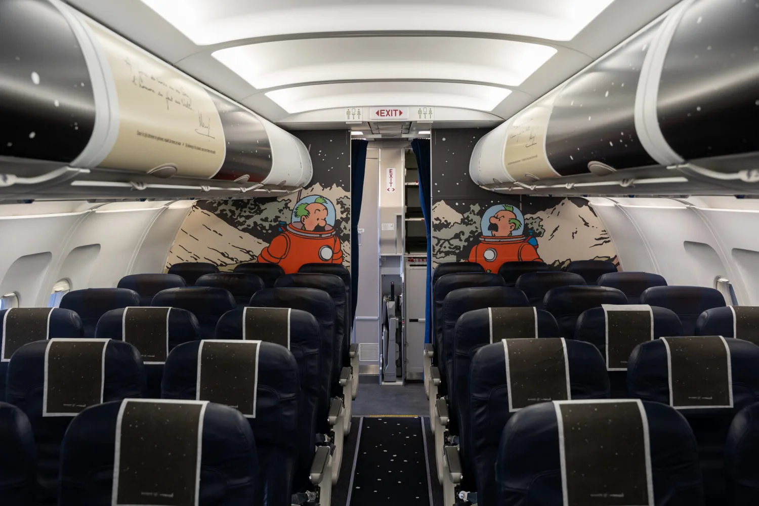 Kabine eines Brussels Airlines Flugzeugs mit Reihen dunkler Passagiersitze und Comic-Weltraumwandgrafik mit Astronautenfiguren und Exit-Schild - © Brussels Airlines / Hergé / Tintinimaginatio 2026