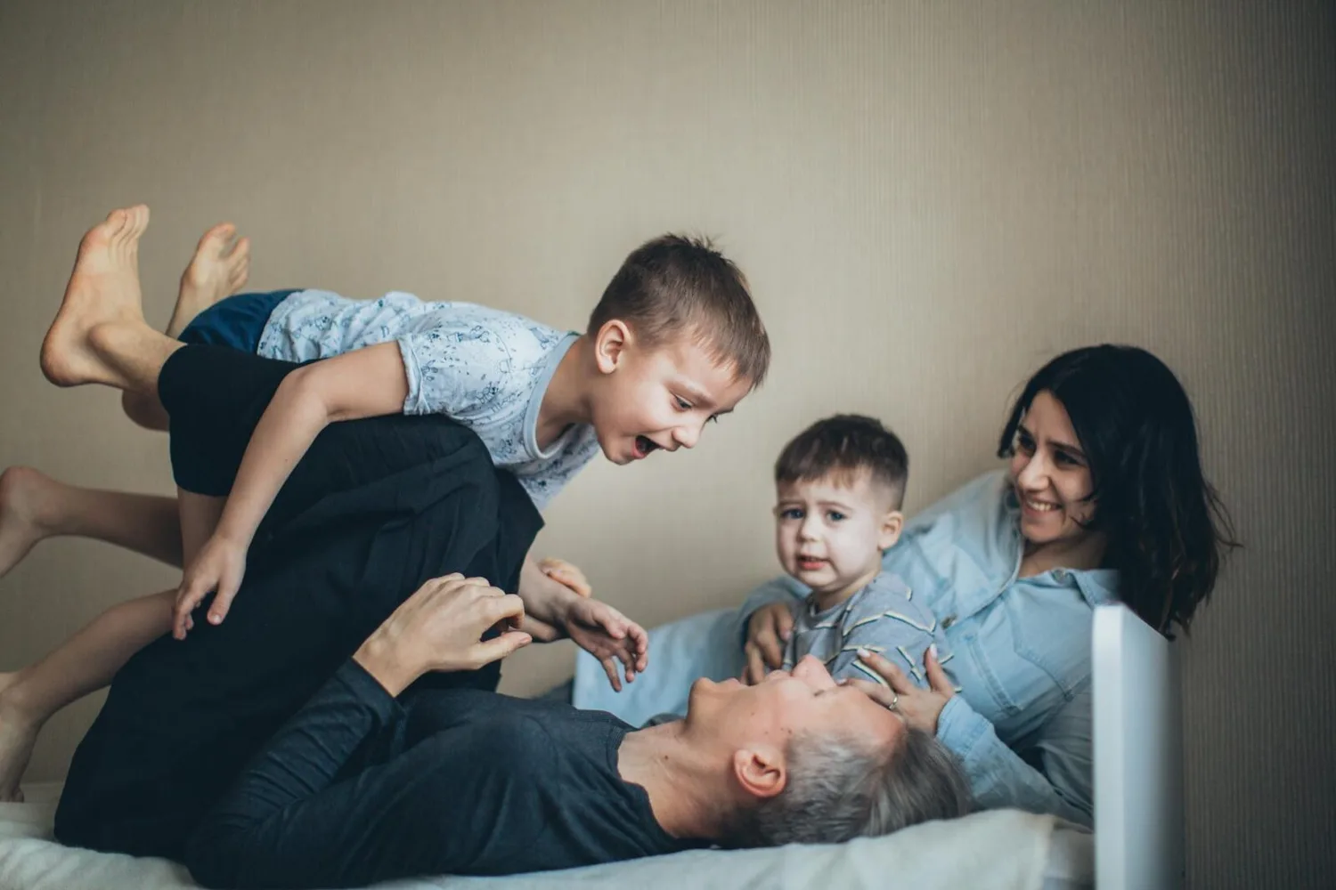 Familie spielt lachend im Bett: Vater hebt älteren Jungen, Mutter hält jüngeren Sohn, freundliche Atmosphäre, Familienzeit, Kita-ähnliche Szene am Flughafen München-Kontext