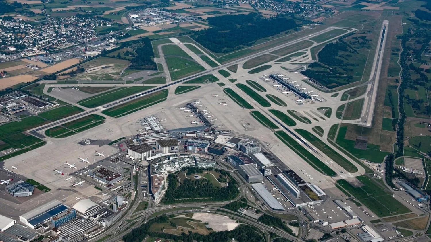 Luftaufnahme Flughafen Zürich mit Terminals, Rollwegen, mehreren startenden und parkenden Verkehrsflugzeugen und umliegender Landschaft (c) Flughafen Zürich