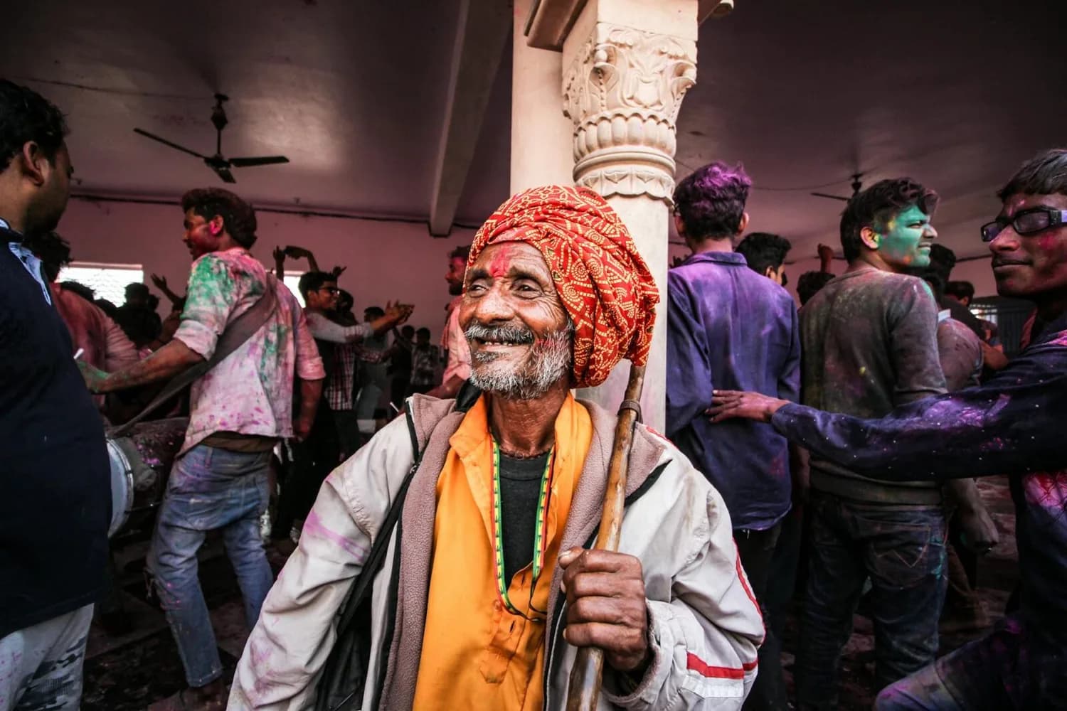 älterer indischer Mann mit rotem Turban und Stock lächelt bei Holi-Fest inmitten feiernder, farbverschmierter Menschen in einer Halle, Symbol für kulturellen Austausch zwischen Air India und Lufthansa in Frankfurt