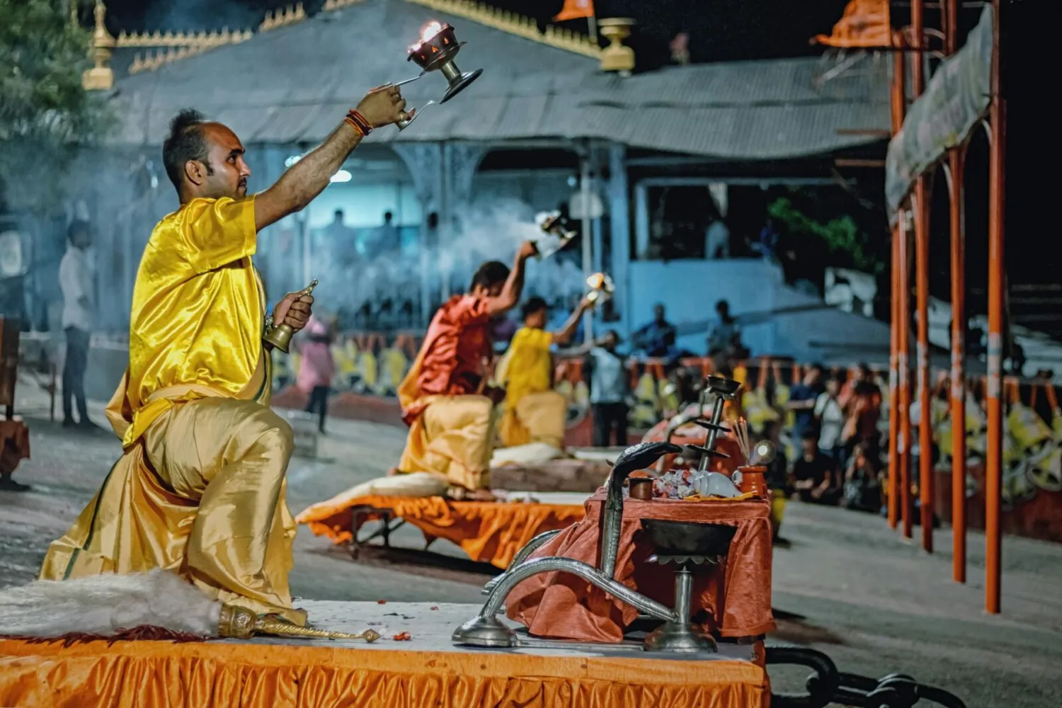 Hindupriester in gelber Kleidung führt abendliche Aarti-Ritual mit brennender Lampe am Flussufer, Hintergrund weitere Priester und Zuschauer — Air India Lufthansa Kooperation erwähnt.