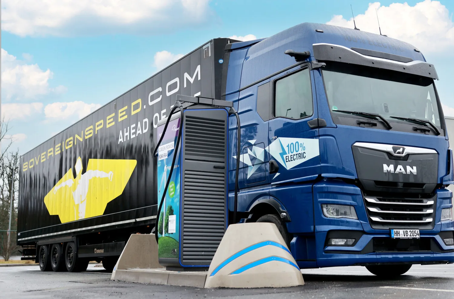 Blauer MAN Elektro-Lkw mit Auflieger und 100% Electric Aufschrift beim Laden an Fraport ePark Lkw-Ladesäule, Parkfläche und Werbegrafik auf der Trailer-Seitenwand