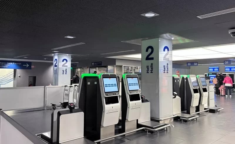 Selbstbedienungs-Check-in-Automaten in Flughafenhalle 2 mit Gepäckwaagen und Reisenden im Hintergrund