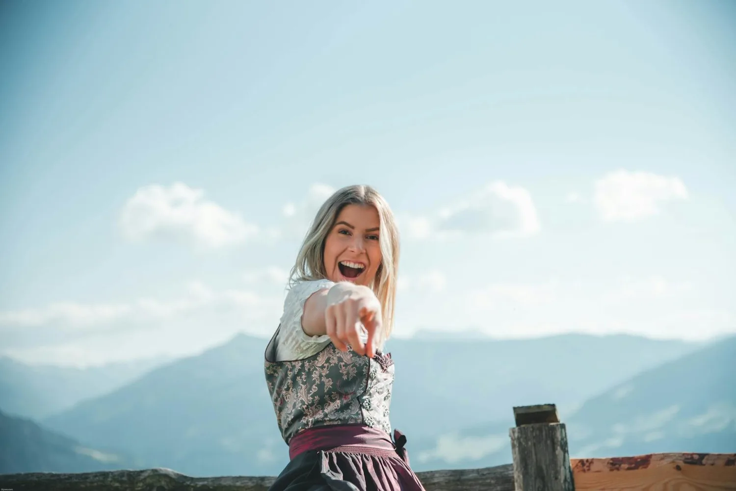 Junge Frau im Dirndl lacht und zeigt mit ausgestrecktem Arm vor Alpenpanorama, bayerische Perspektive FSJ