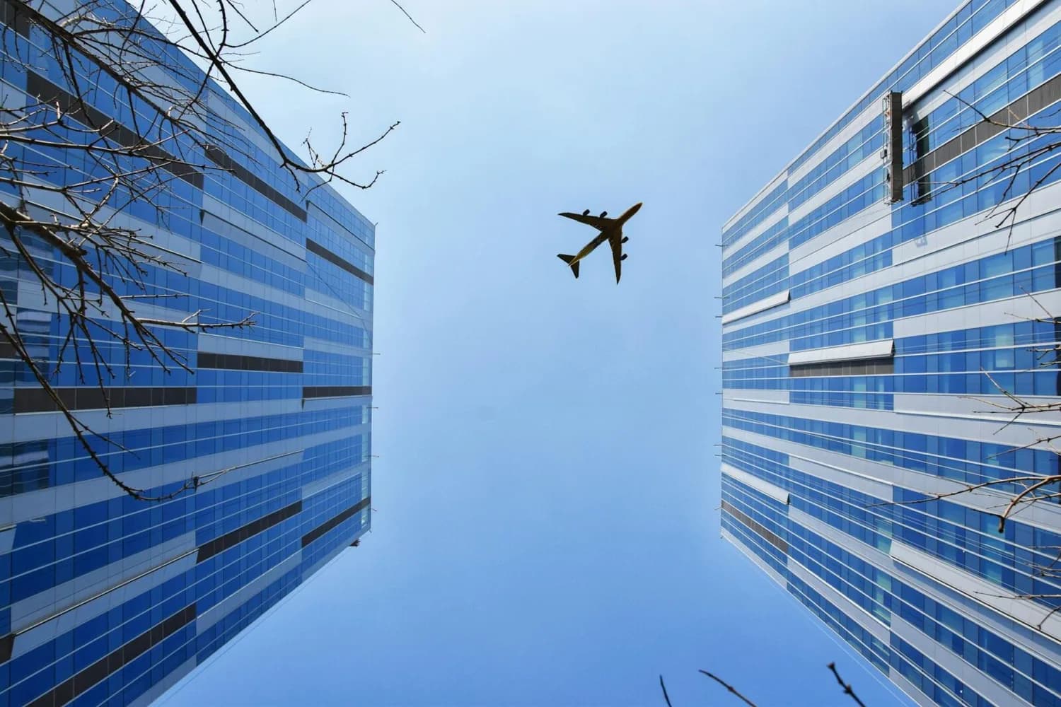 Flugzeug fliegt über städtische Hochhäuser bei klarem Himmel, Blick von unten zwischen Glasfassaden, Schiphol leisere Flugzeuge.