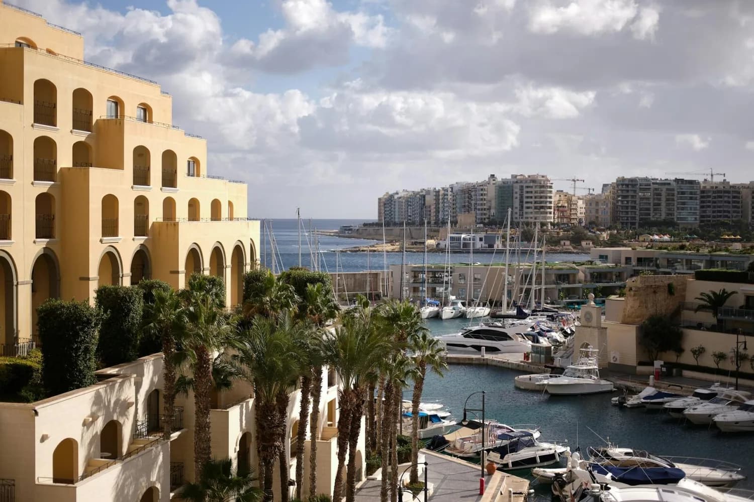 Hafen mit Yachten und Segelbooten, gelbes mediterranes Hotel mit Arkaden und Palmen, Blick auf Küstenpromenade und Wohnhochhäuser in Malta (Flugroute Brüssel–Malta)