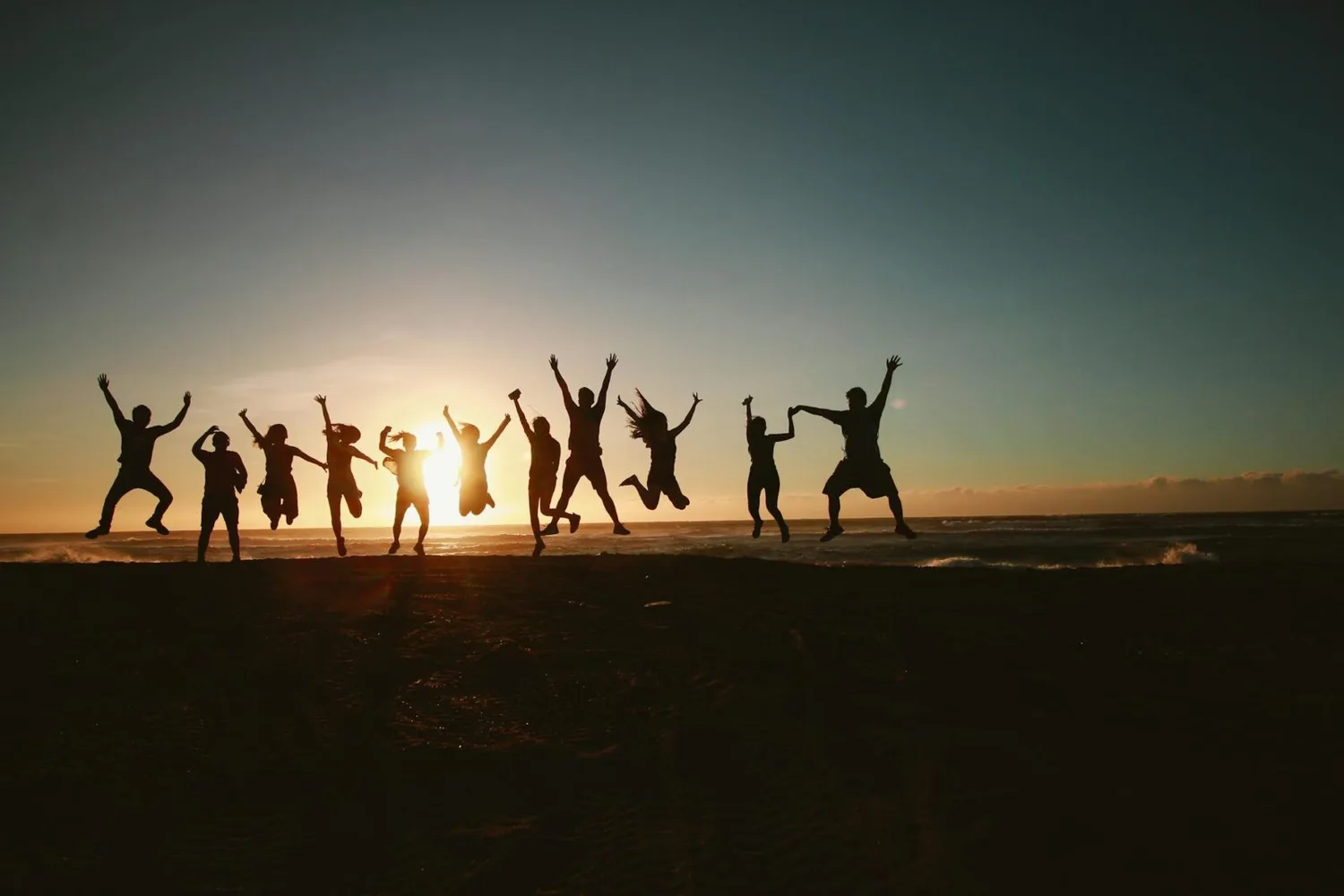 Gruppe junger Menschen springt am Strand bei Sonnenuntergang, fröhlicher Strandurlaub nahe Nantes, Silhouetten vor Meer und Himmel