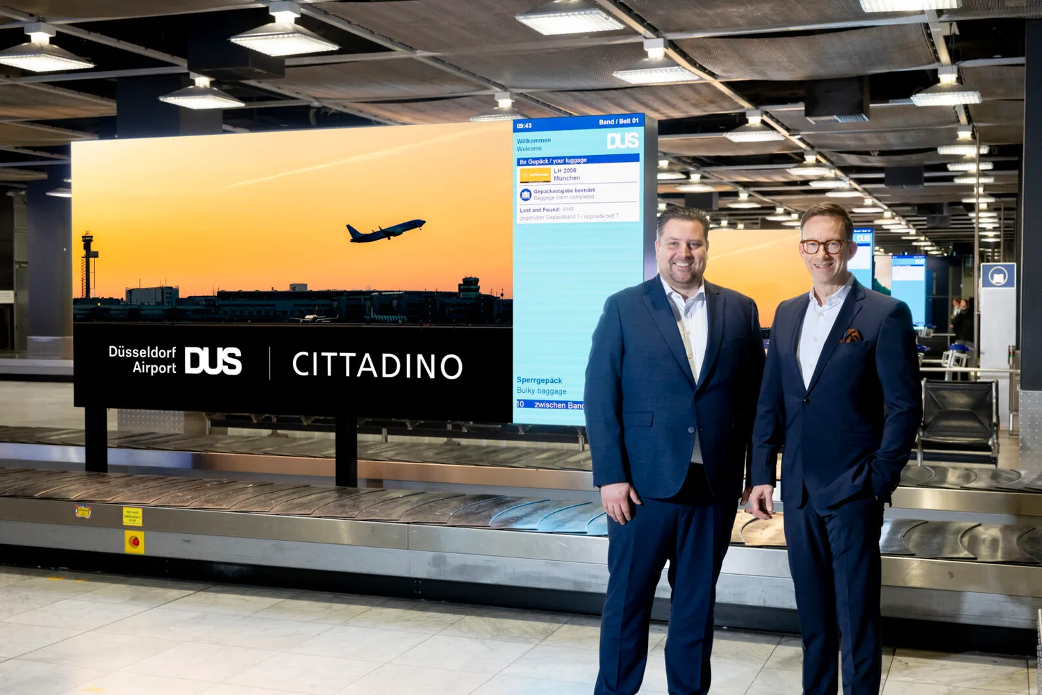 Zwei Geschäftsleute vor großem DUS Welcome Board am Gepäckband im Flughafen Düsseldorf mit Flugzeug-Silhouette und Cittadino-Logo