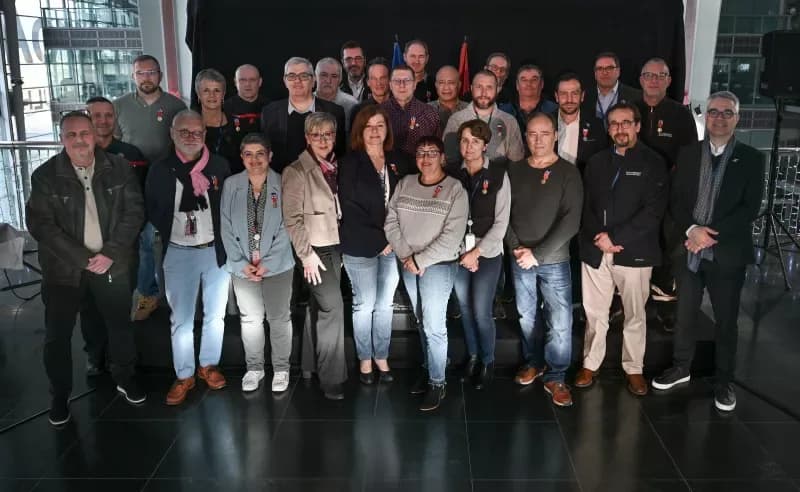 Gruppenfoto mehrerer Männer und Frauen in Innenraum bei Verleihung mit am Revers sichtbaren Medaillen und Flaggen im Hintergrund