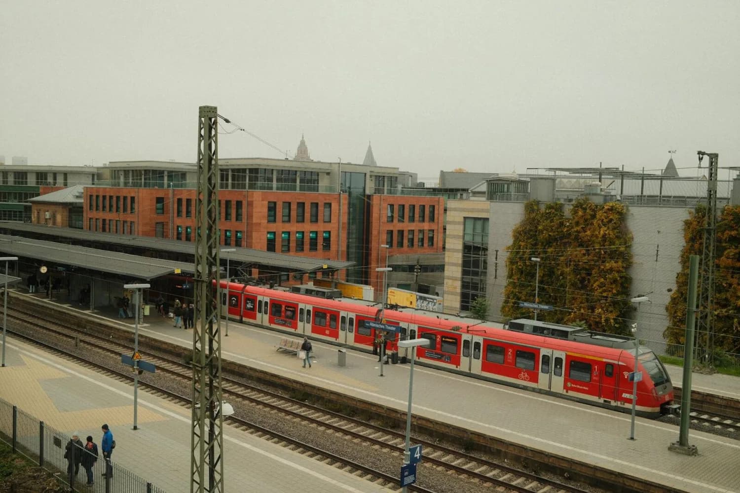 Mainz Hauptbahnhof Bahnsteig mit roter S‑Bahn, wartenden Fahrgästen und modernen Bürogebäuden im Hintergrund