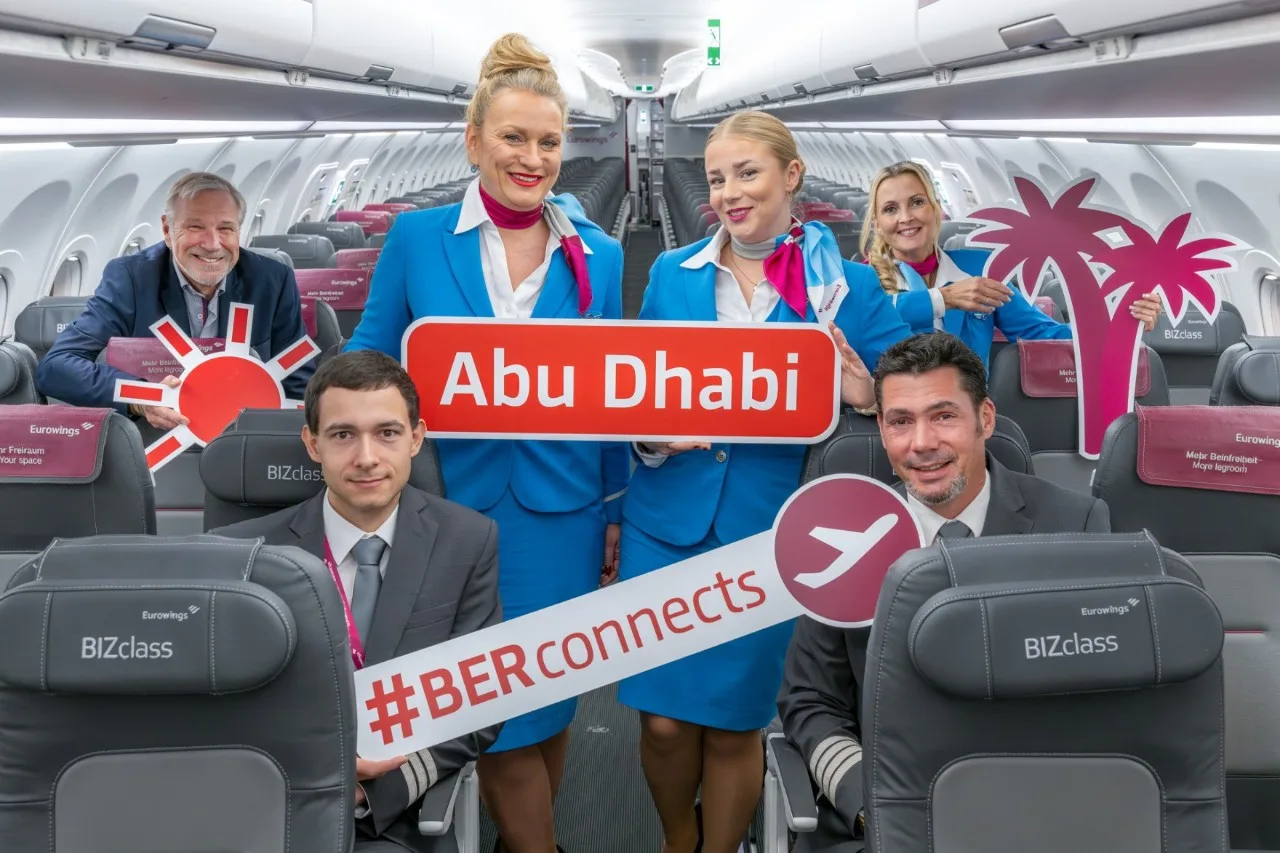 Flugpersonal und Piloten in Eurowings BIZclass Kabine halten Schilder Abu Dhabi und #BERconnects, Werbemotiv für Direktverbindung mit Palmen- und Sonnen-Icons