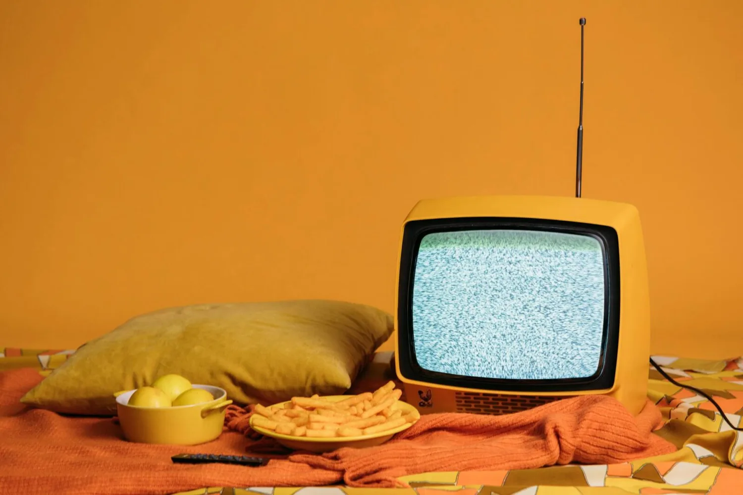 Gelber Retro-Fernseher mit Bildstörung neben Teller mit Snacks, Schüssel mit Zitronen und Kissen vor orangefarbenem Hintergrund, Serie Amsterdam Schiphol