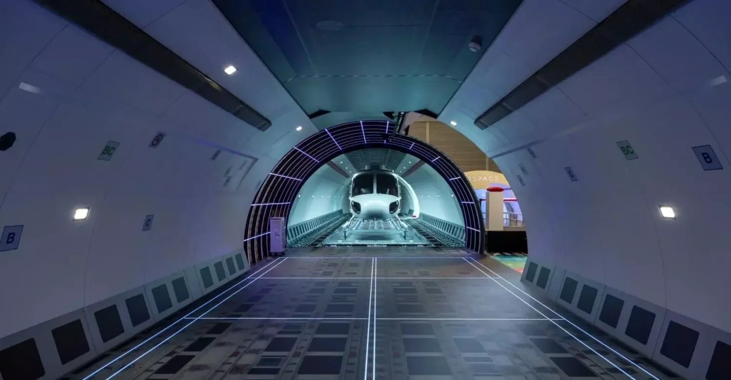 Innenansicht eines Airbus-Frachterrumpfs mit Ladungsschiene und einem vorderen Flugzeugrumpf im Laderaum, futuristische Beleuchtung und Markierungen auf dem Boden