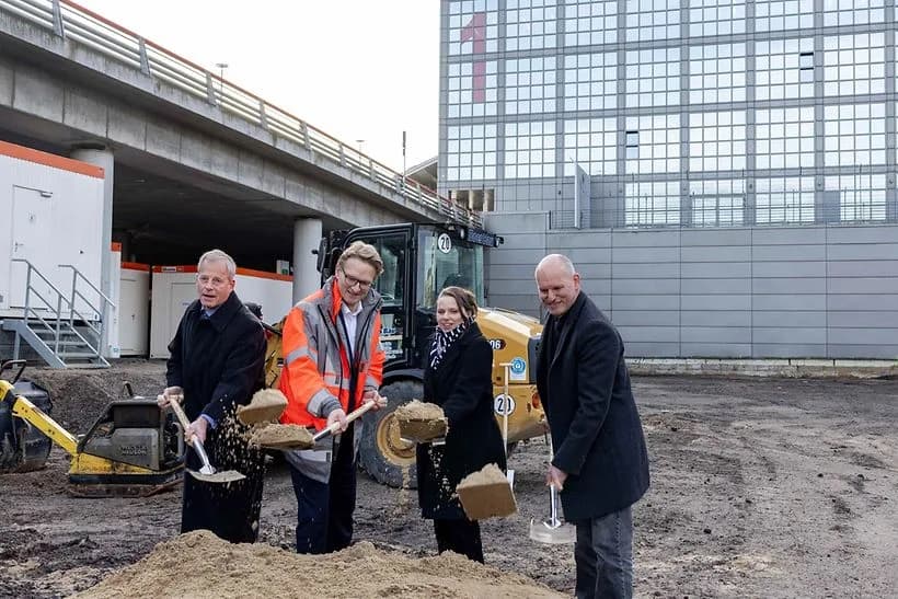 Symbolischer Spatenstich auf Baustelle für neue Sortierhalle mit vier Personen, Schaufeln und Baumaschine im Hintergrund