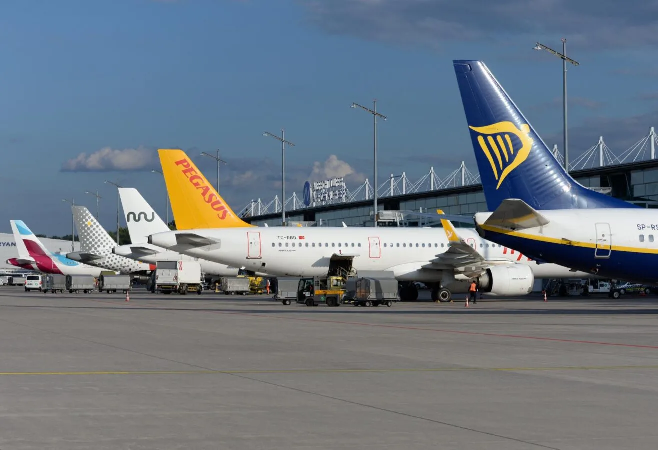 Reihe geparkter Passagierflugzeuge mit sichtbaren Leitwerken verschiedener Airlines (Ryanair, Pegasus, weitere) am Flughafen neben Terminal und Bodenfahrzeugen