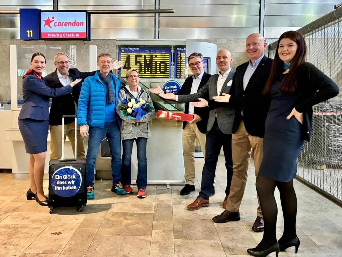 Gruppenfoto am Flughafen Nürnberg vor dem Corendon‑Priority‑Check‑in. Eine Gruppe von Mitarbeitenden und einem Reisendenpaar steht lächelnd zusammen. Die zentrale Person hält einen Blumenstrauß, daneben wird ein Modellflugzeug von Corendon präsentiert. Im Vordergrund steht ein schwarzer Koffer mit dem Aufdruck ‚Ein Glück, dass wir ihn haben‘. Im Hintergrund sind Anzeigetafeln für Abflüge zu sehen. 4,5-Millionster Fluggast: Petra Beer (4. v.l.) mit Waldemar Strzelczyk (3. v.l.) aus der Nähe von Erlangen wurde von Flughafengeschäftsführer Dr. Michael Hupe (2. v.l.), COO Jan C. Bruns (2. v.r.), Leiter Aviation Vertrieb Christian Käser (3. v.r.) und Christian Hein, Head of Sales & Marketing von Corendon Airlines (4. v.r.), überrascht. Corendon-Cabin-Crew-Mitglied Anna (links) und Schichtleiterin Ricarda Ley (rechts) gratulieren ebenfalls. Foto: Christian Albrecht / Airport Nürnberg