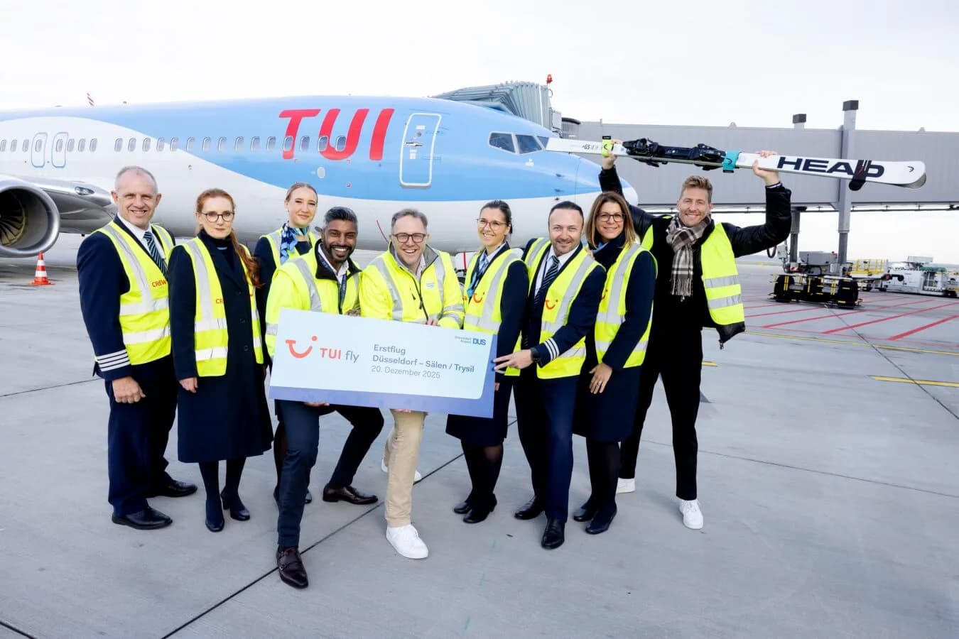 TUI fly Crew und Flughafenpersonal mit Sicherheitswesten vor TUI-Flugzeug am Gate, halten Willkommenscheck und Skier für Erstflug Düsseldorf–Sälen/Trysil