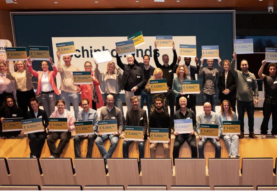 Gruppenfoto von Gewinnern bei Preisverleihung am Flughafen Schiphol, Personen halten Schecks mit Förderbeträgen und Schriftzug Schipholfonds im Hintergrund
