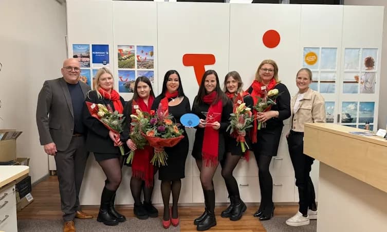 Reisebüro-Team im TUI Center am Flughafen Paderborn, mehrere Mitarbeiterinnen mit roten Schals halten Blumensträuße und Auszeichnungen vor Firmenlogo