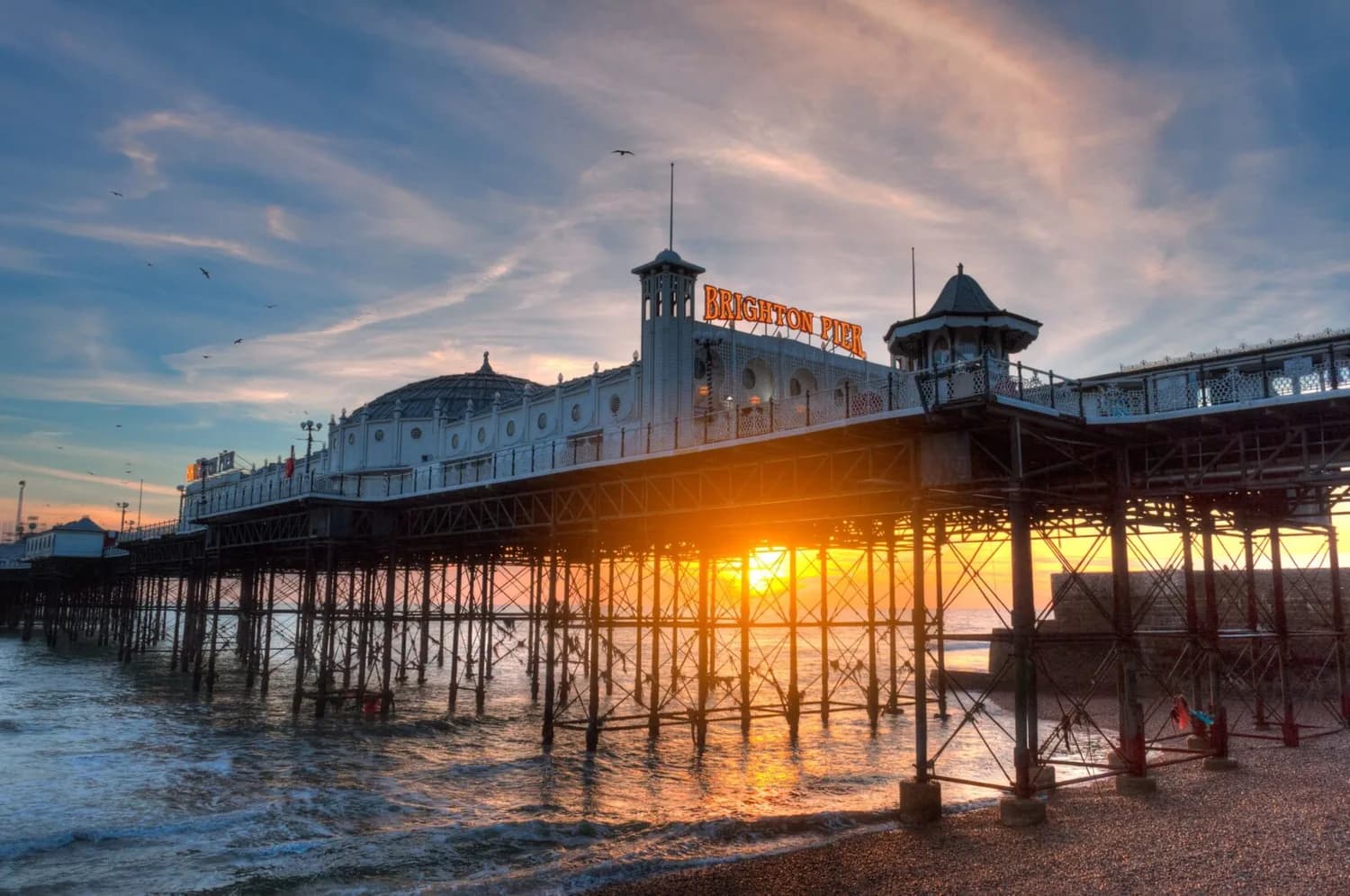 Brighton Pier bei Sonnenuntergang mit leuchtendem Schriftzug, Eisenkonstruktion über dem Meer und spiegelndem Wasser am Kiesstrand
