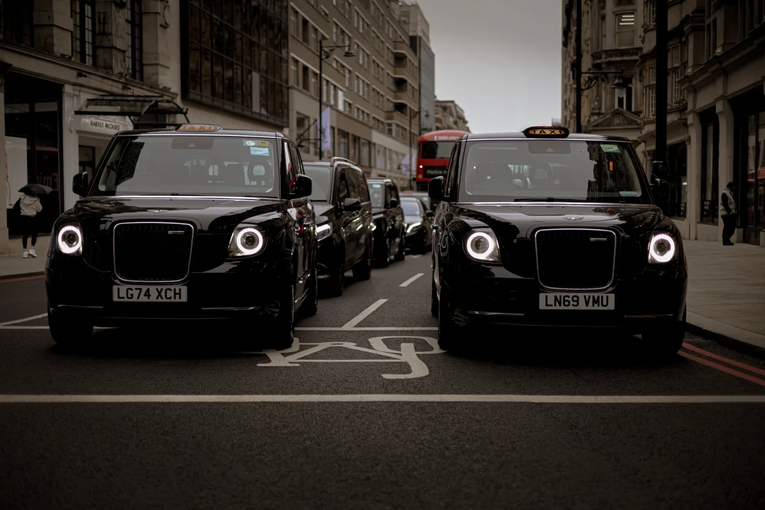 Zwei schwarze London-Taxis nebeneinander im Stadtverkehr vor Gebäuden mit Doppeldeckerbus und wartenden Fahrzeugen