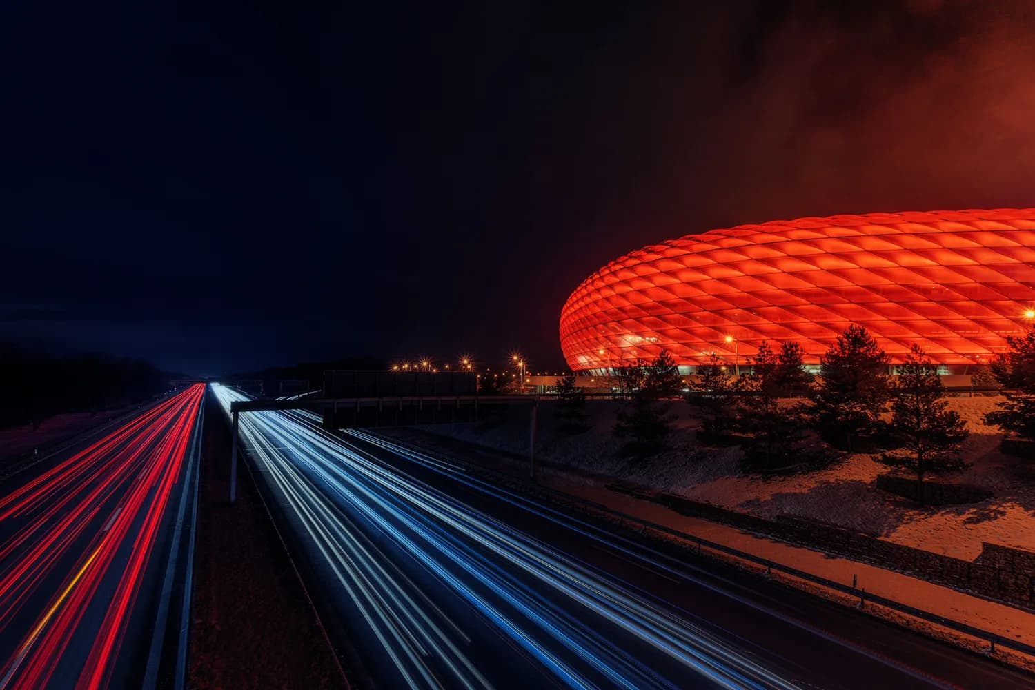Allianz Arena in München nachts rot beleuchtet neben Autobahn mit langen Lichtspuren von Fahrzeugen