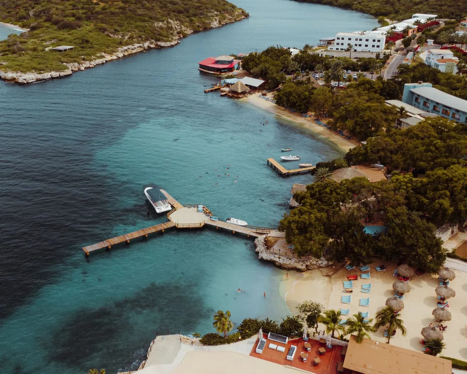 Luftaufnahme einer karibischen Bucht auf Curaçao mit Holzsteg, Booten, türkisfarbenem Meer, Sandstrand, Sonnenschirmen und Hotelanlagen