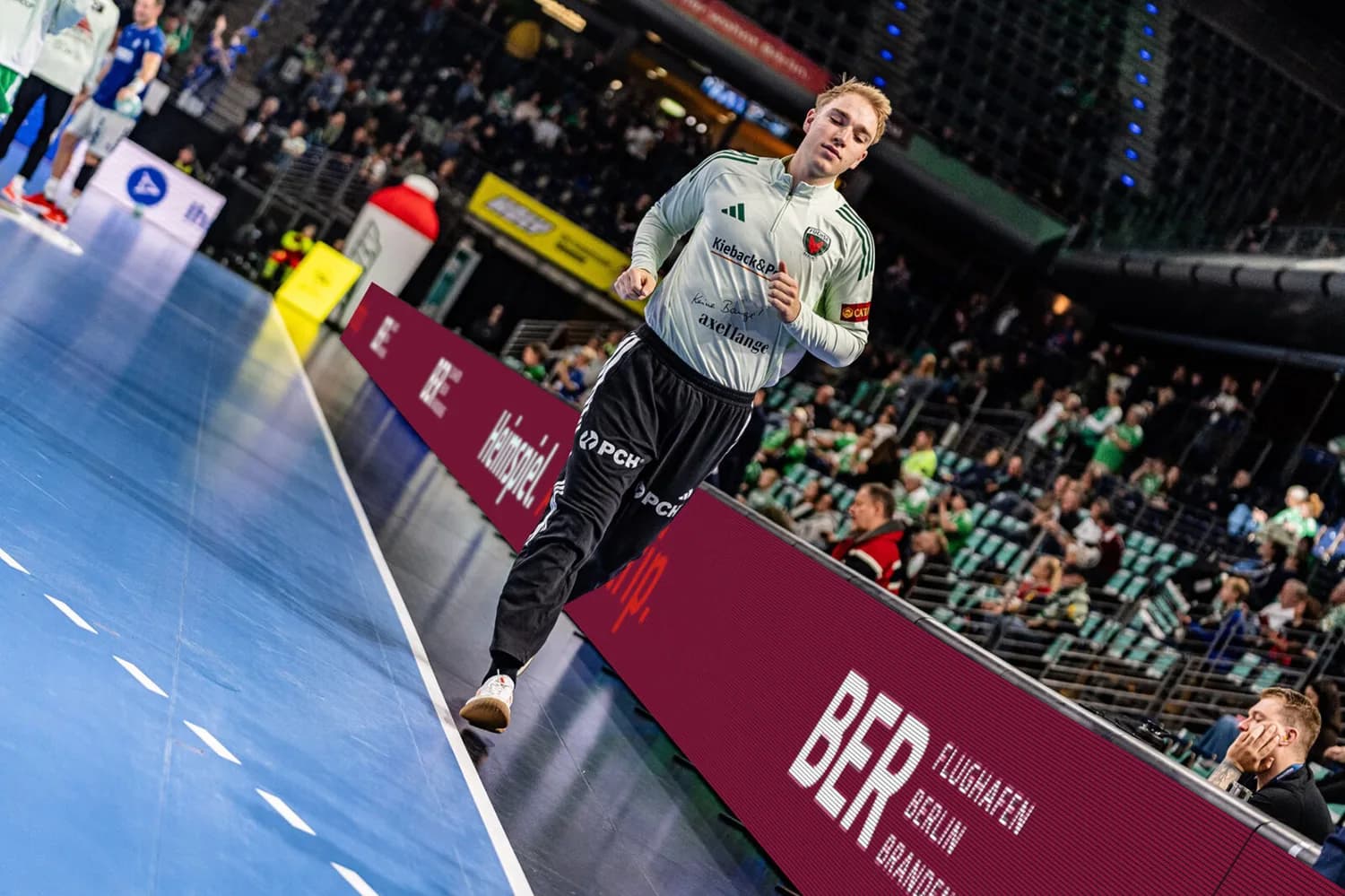 Handballtorwart der Füchse Berlin joggt in Aufwärmshirt und Trainingshose am Spielfeldrand vor Zuschauern und Werbebande