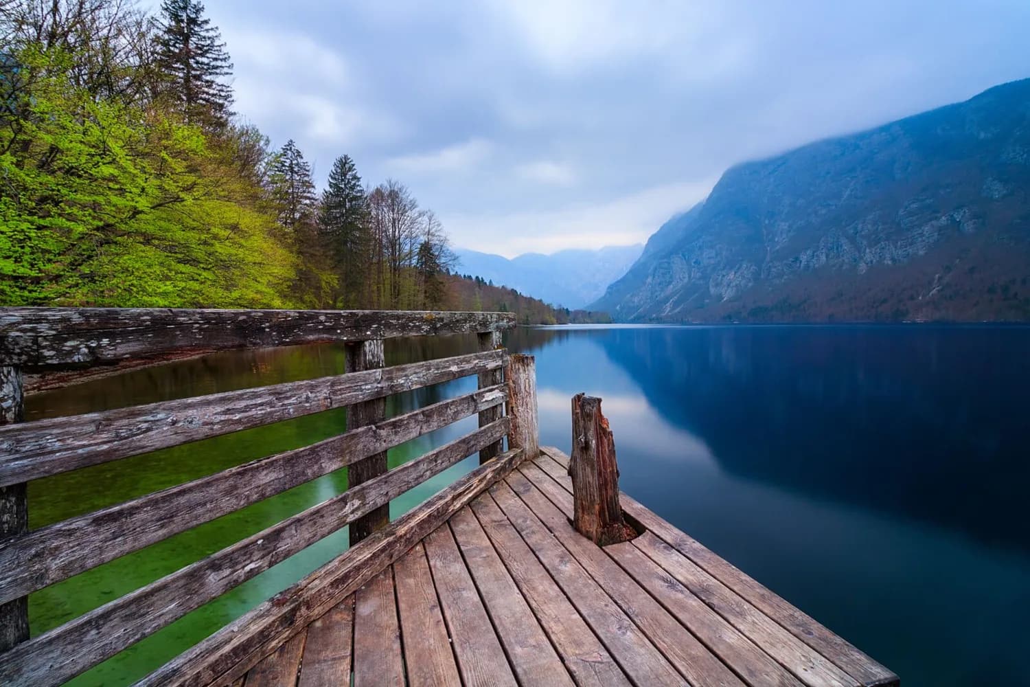 Holzsteg mit verwittertem Geländer am smaragdgrünen Bohinjsee in Slowenien, ruhige Bergsee-Reflexionen und bewaldete Ufer bei dunstigem Himmel