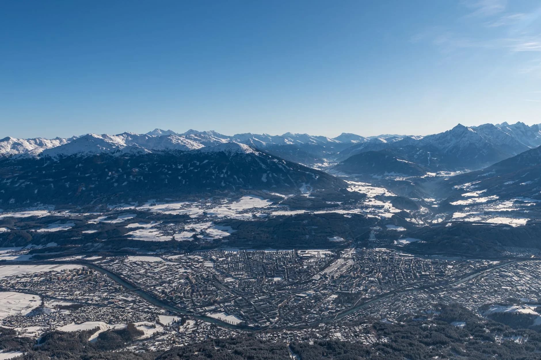 Luftaufnahme eines verschneiten Alpentals mit Stadt am Flussufer, umgeben von schneebedeckten Berggipfeln