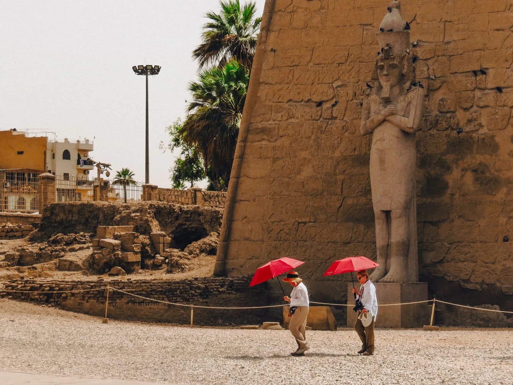Zwei Touristen mit roten Sonnenschirmen gehen an der Pylonstatue des Luxor-Tempels in Ägypten vorbei
