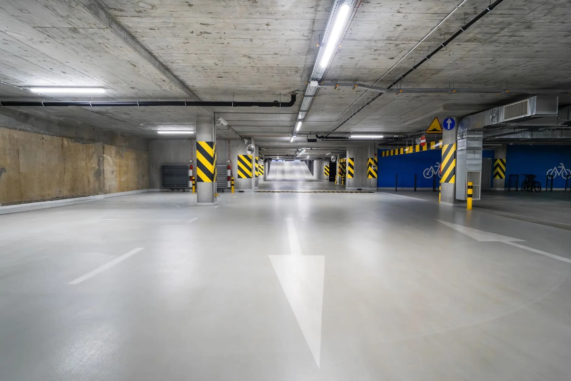 Leeres Parkdeck mit markierten Fahrbahnpfeilen, gelb-schwarzen Stützpfeilern und Fahrradständern an blauer Wand
