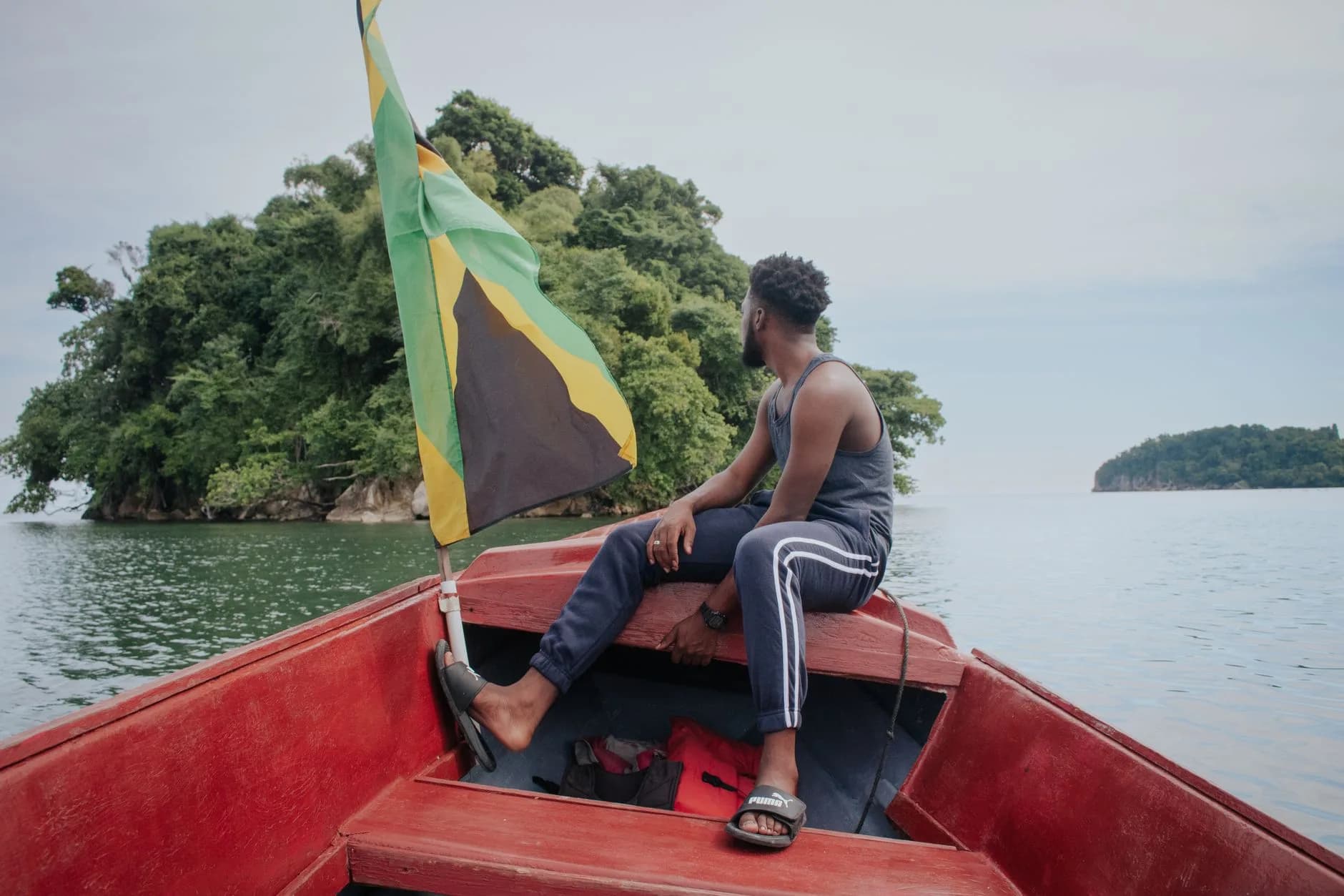 Mann sitzt im Bug eines roten Bootes mit jamaikanischer Flagge, Blick auf bewaldete Inseln im Wasser