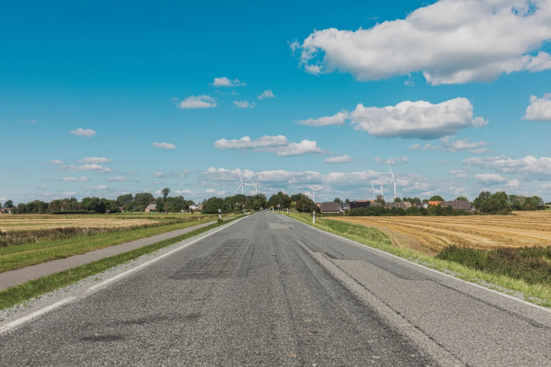 Leere Landstraße führt durch abgeerntete Felder auf ein Dorf mit Windrädern unter blauem Himmel