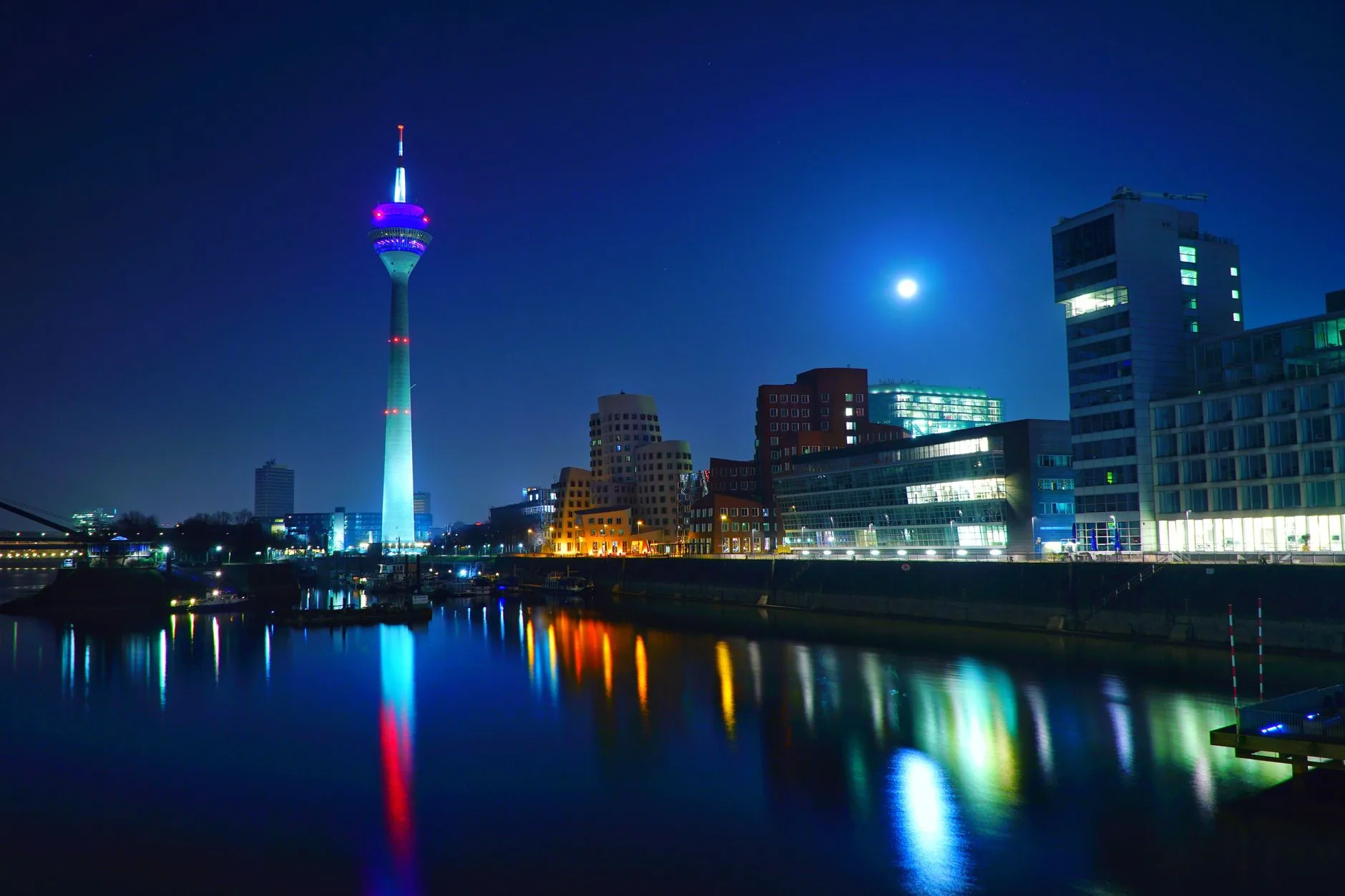 Beleuchteter Fernsehturm und Hafengebäude bei Nacht spiegeln sich im ruhigen Wasser des Medienhafens