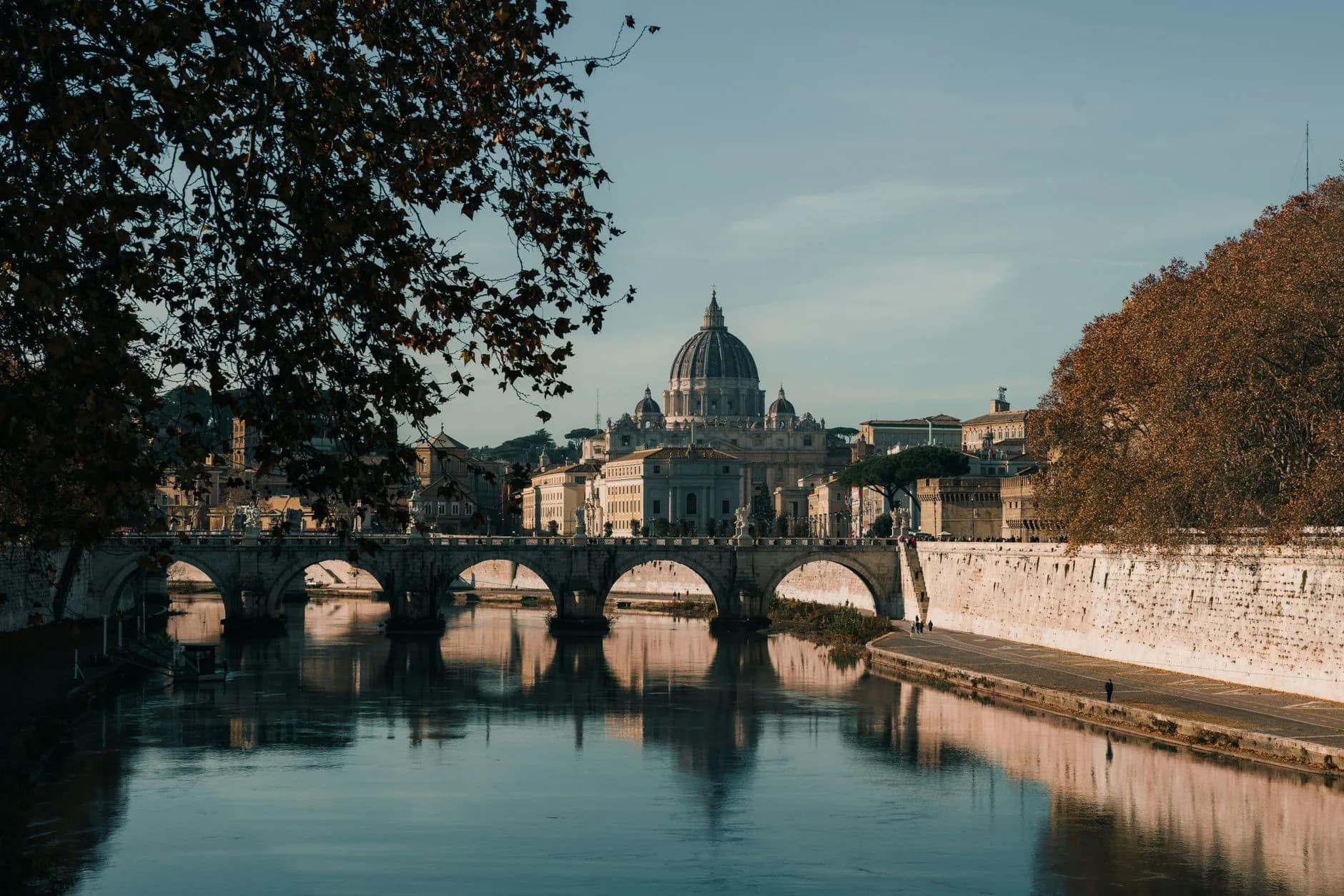 Blick über den Tiber auf eine Bogenbrücke mit der Kuppel des Petersdoms im Hintergrund