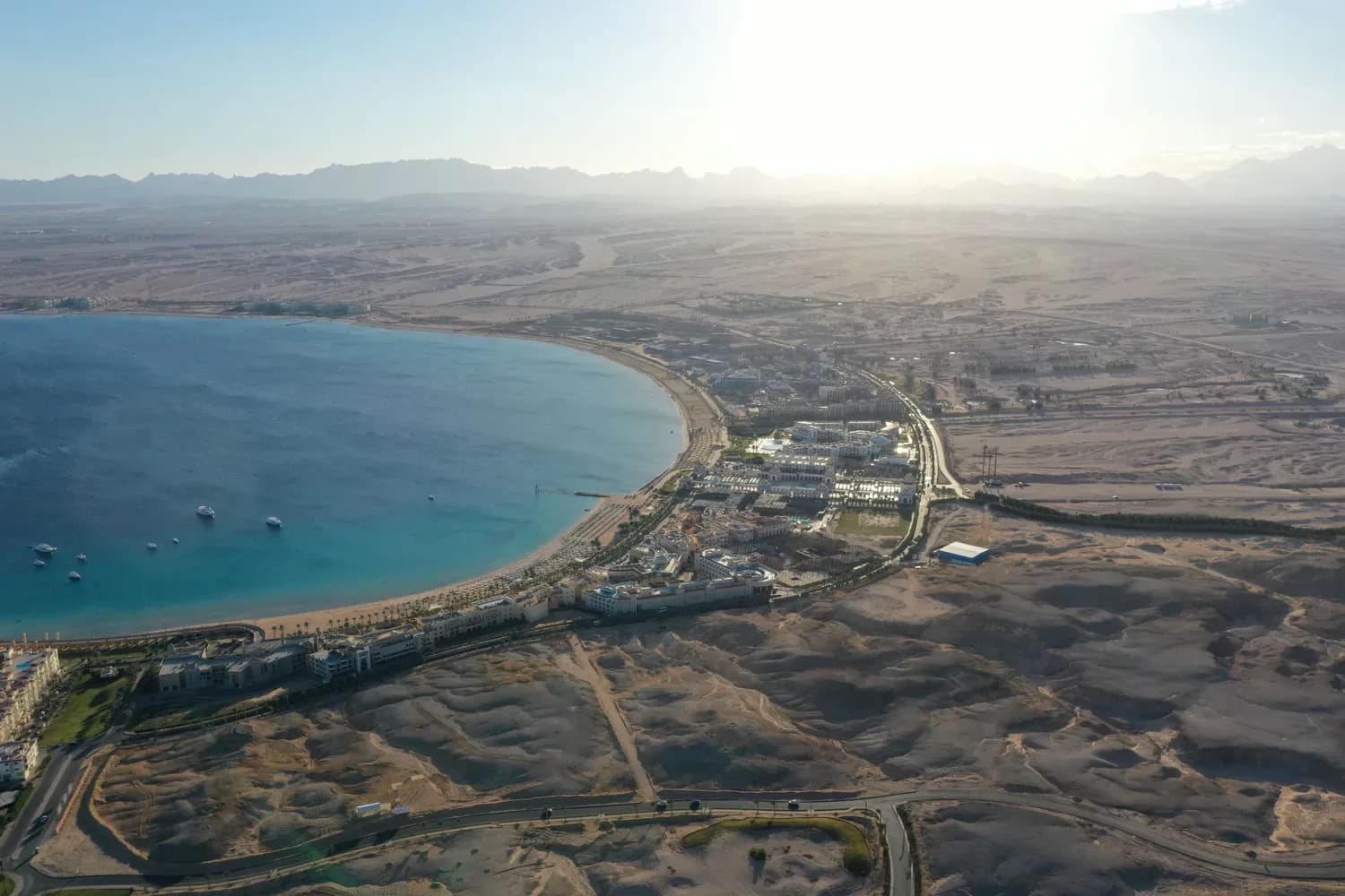 Luftaufnahme einer Bucht mit türkisem Wasser, Hotelanlagen am Strand und Wüstenlandschaft im Hintergrund