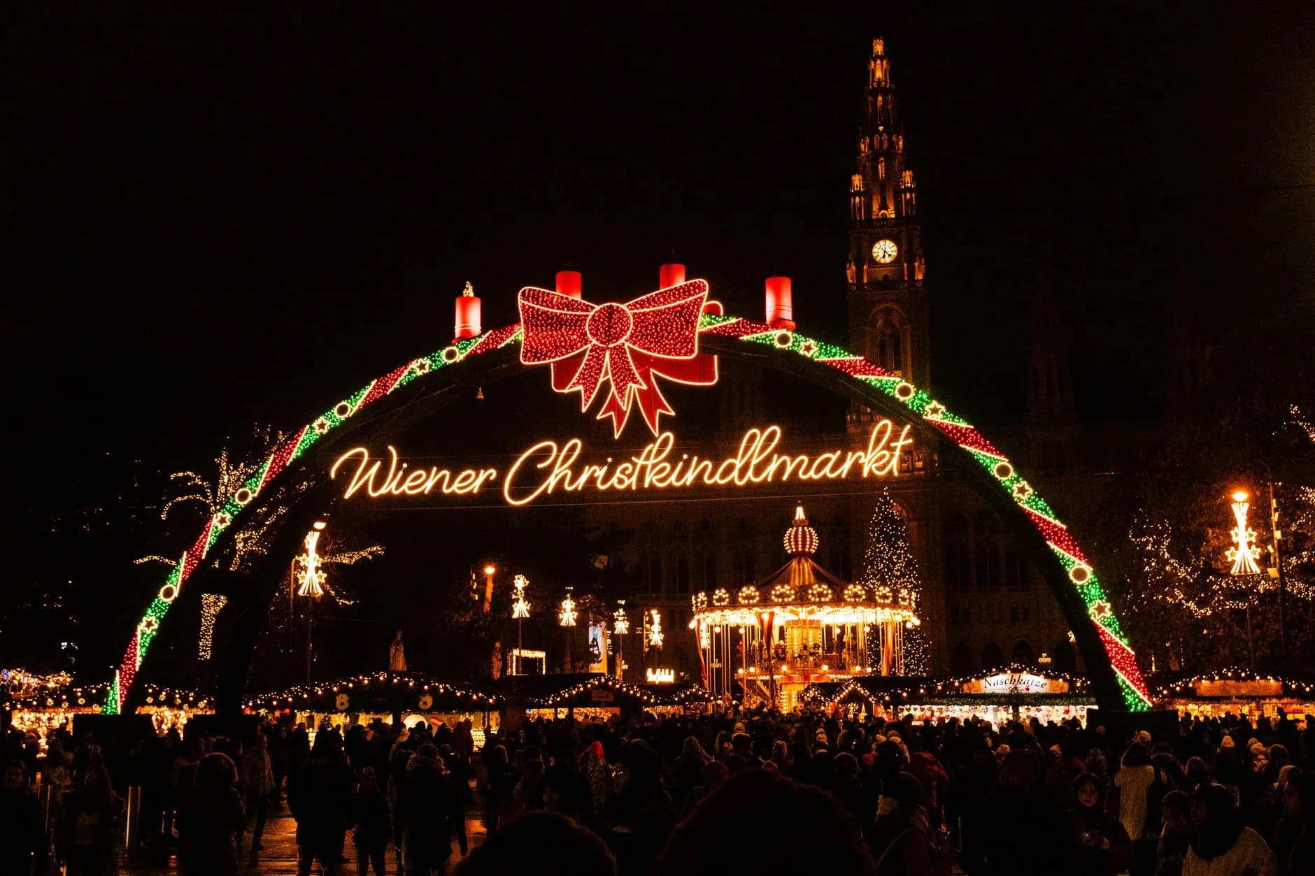 Beleuchteter Eingangsbogen zum Wiener Christkindlmarkt bei Nacht mit Rathaus und Menschenmenge im Hintergrund