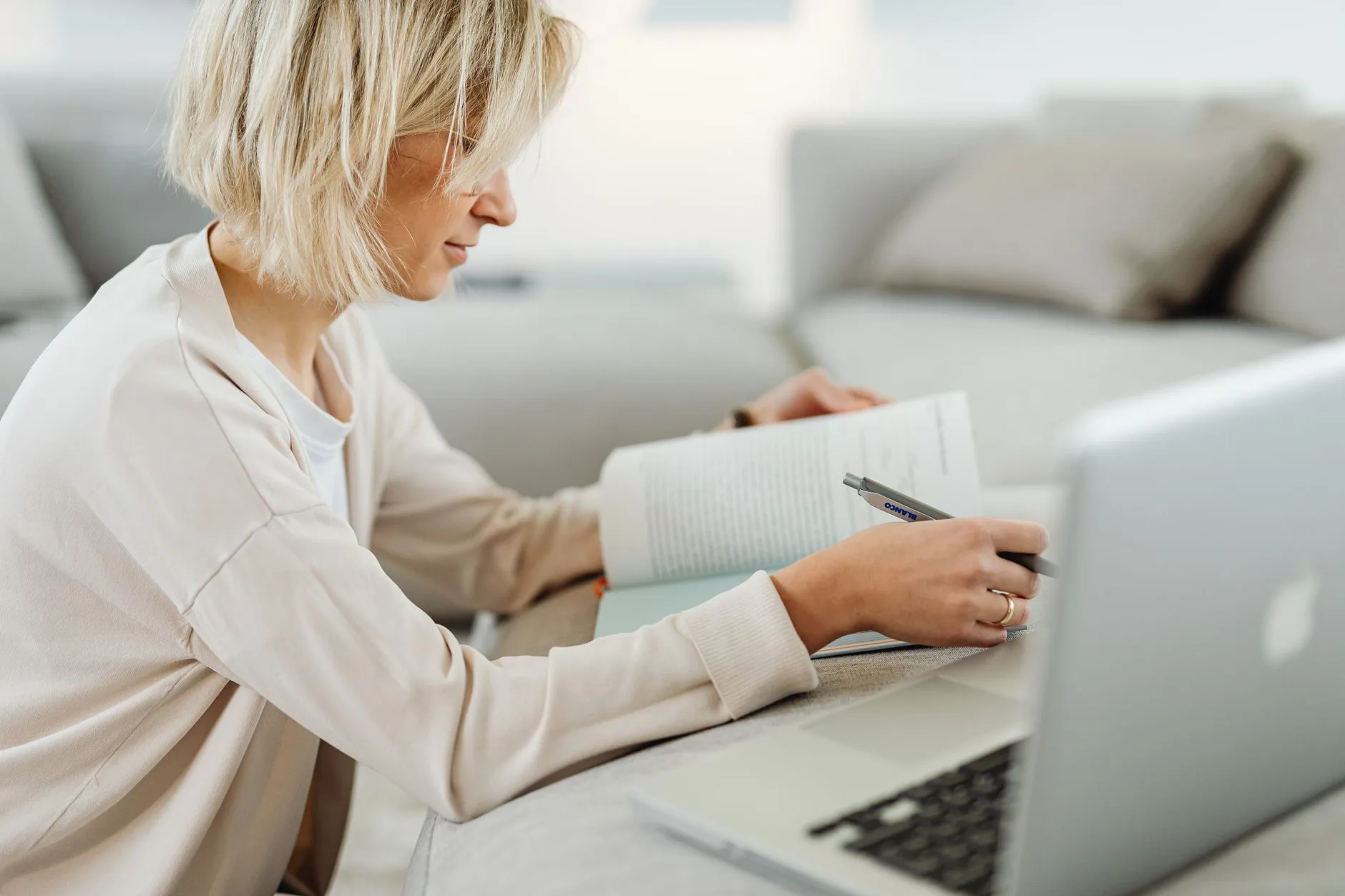 Frau mit kurzen blonden Haaren macht sich Notizen in einem Heft vor einem geöffneten Laptop