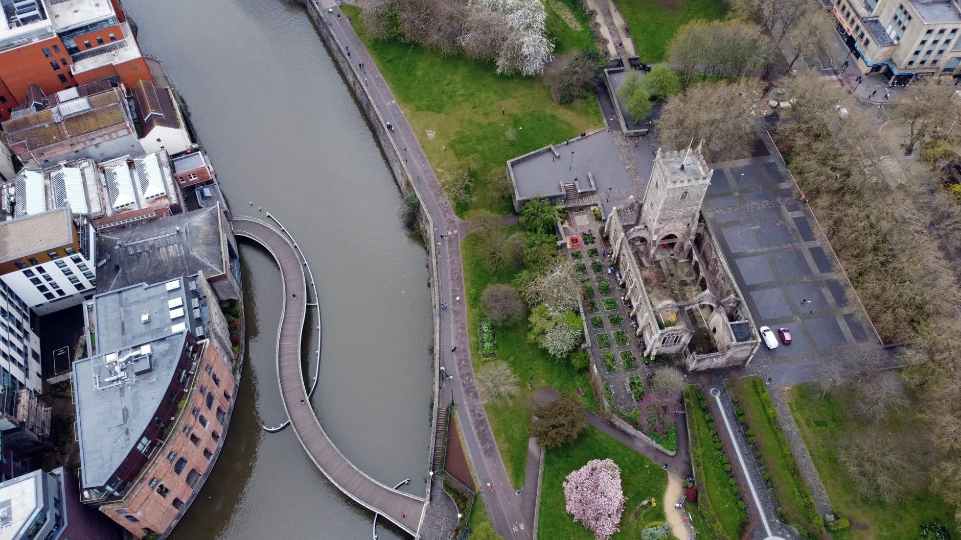 Luftaufnahme einer geschwungenen Fußgängerbrücke über einen Fluss mit angrenzendem Park und Kirchenruine