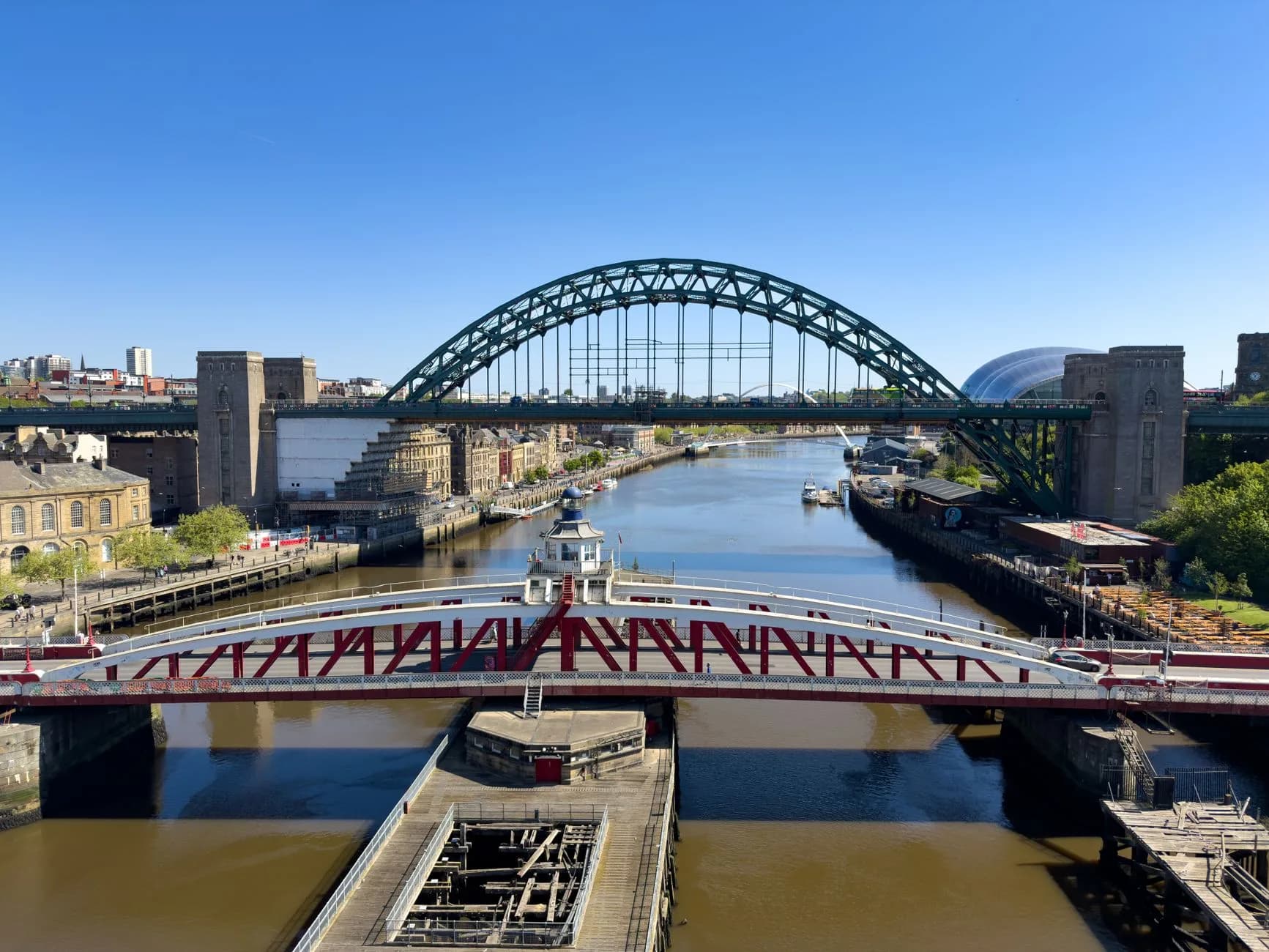 Blick auf den Fluss Tyne mit roter Drehbrücke im Vordergrund und grünem Brückenbogen im Hintergrund bei klarem Himmel